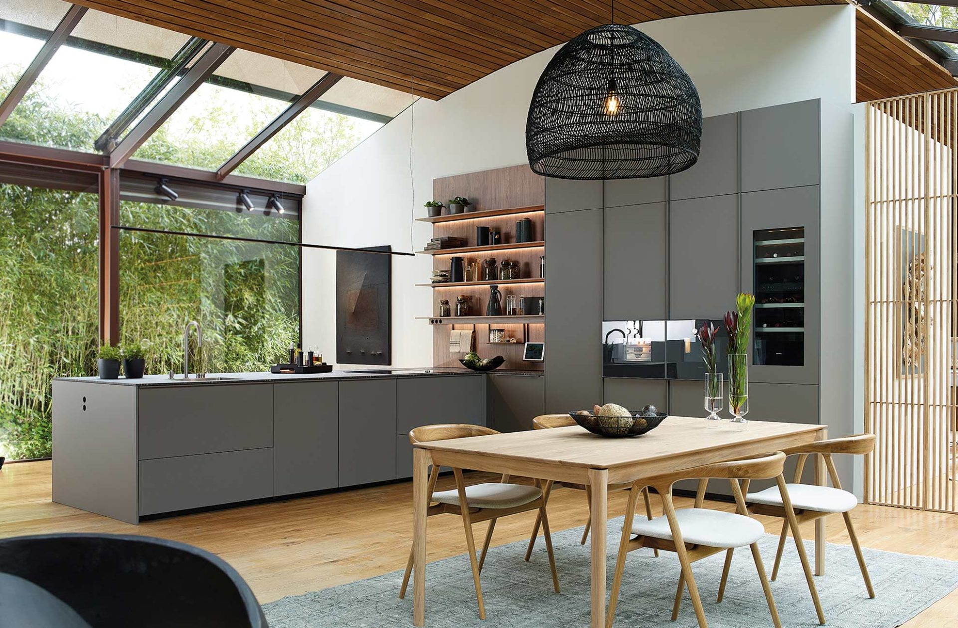 Modern slate grey peninsula kitchen next to a wooden dining table and enclosed by floor-to-ceiling glazing.