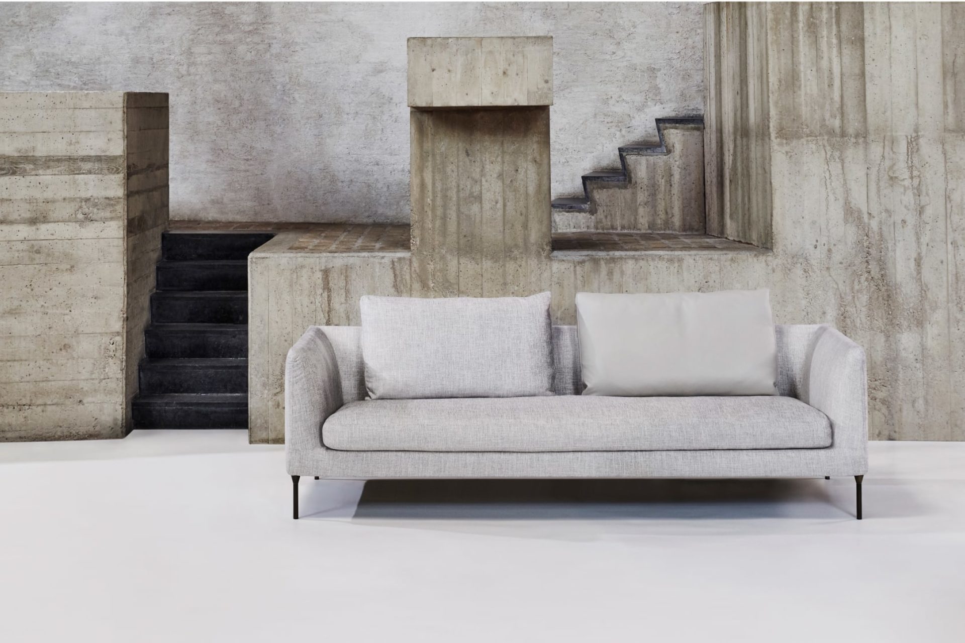 Sofá minimalista de diseño escandinavo con estructura rectangular en tejido gris claro y patas en acero negro, modelo Delta de Bensen, disponible en eba